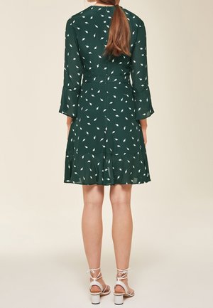 Vrouw met lang bruin haar, gekleed in een groene jurk met witte bladmotieven en witte sandaalhakken, staand met de rug naar een beige achtergrond.