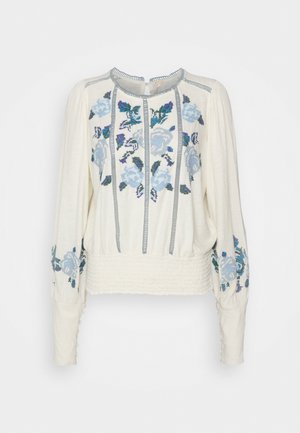 Blusa de manga larga color crema con cintura fruncida, puños abotonados, bordado floral azul y púrpura en el frente y las mangas, y detalles en ribete gris.