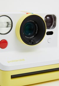 Polaroid camera featuring a white body with yellow accents, round lens, red shutter button, and a colorful stripe on top. Filter sensor included.