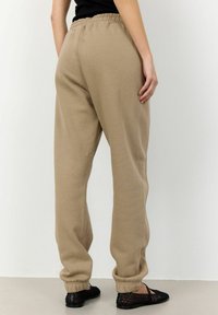 Beige Jogginghosen mit einem elastischen Bund und umgeschlagenen Säumen, die eine glatte Textur und einen lässigen Schnitt aufweisen, kombiniert mit schwarzen geflochtenen Schuhen.