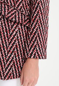 Detaljebillede af en hånd ved siden af en uldfrakkeærme med røde, sorte og hvide zigzag-mønstre og en detalje med bæltestrop i stof.