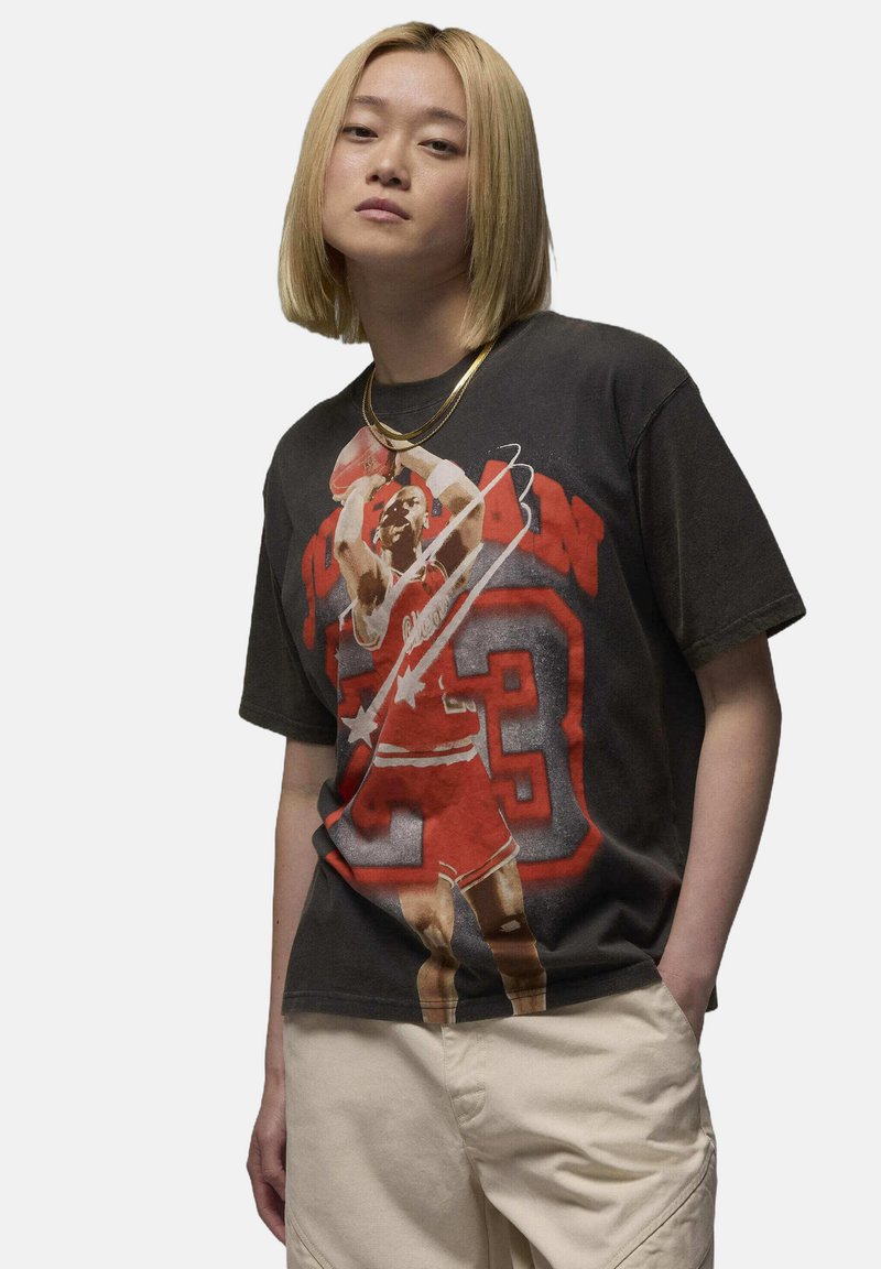 Jeune personne aux cheveux blonds mi-longs portant un t-shirt graphique de basket-ball noir de Michael Jordan et un pantalon beige clair, se tenant avec une main dans la poche.