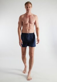 Marineblauwe boxers met een subtiel patroon, elastische tailleband en losse pasvorm, gedragen door een model tegen een simpele witte achtergrond.