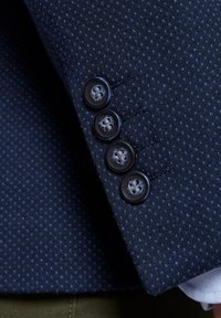 Granatowy teksturowany materiał garniturowy z drobnym wzorem w kropki, z czterema ciemnymi guzikami na mankietach i szczegółowym przeszyciem.
