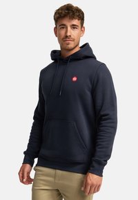 Bărbat purtând un hanorac bleumarin cu un logo circular roșu pe piept și pantaloni bej, stând pe un fundal alb simplu.