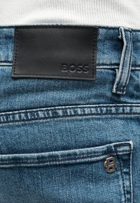 Jean en denim bleu avec une étiquette en cuir noir embossée « BOSS » et un petit logo métallique sur la poche arrière.