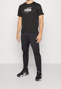 Man i svart Nike t-shirt, svarta sportiga joggare och svarta Nike-sneakers som står framför en enfärgad bakgrund.