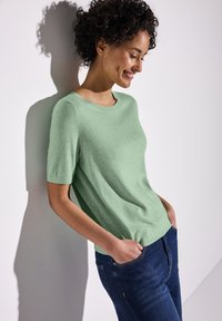 Pull à manches courtes de couleur vert clair, fabriqué dans un matériau doux, avec un col rond et une coupe décontractée, associé à un jean en denim foncé.