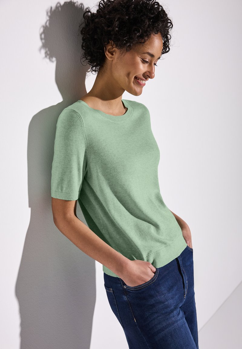 Pull à manches courtes de couleur vert clair, fabriqué dans un matériau doux, avec un col rond et une coupe décontractée, associé à un jean en denim foncé.
