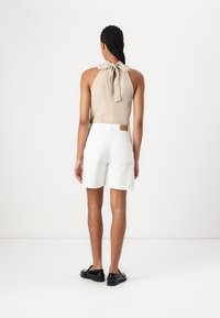Top halter beige con lazo en el cuello, combinado con pantalones cortos blancos con bolsillos traseros y una etiqueta de cuero marrón. Unos zapatos negros completan el look.