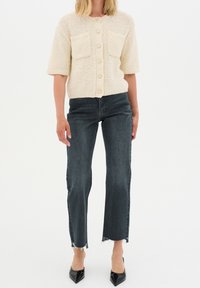 Cárdigan de punto color crema con mangas cortas y dos bolsillos frontales, combinado con jeans oscuros de tiro alto con dobladillos deshilachados y tacones negros puntiagudos.