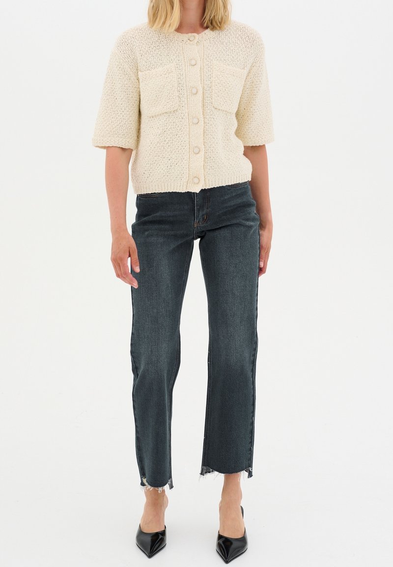Cárdigan de punto color crema con mangas cortas y dos bolsillos frontales, combinado con jeans oscuros de tiro alto con dobladillos deshilachados y tacones negros puntiagudos.