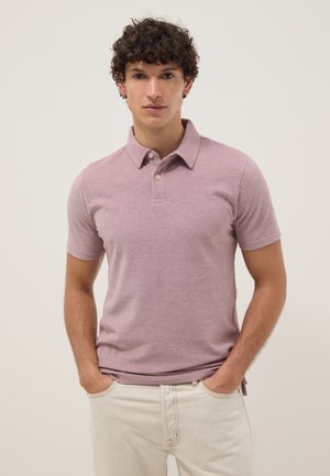 Jeune homme aux cheveux bouclés portant un polo à manches courtes violet clair et un pantalon beige clair, debout les mains dans les poches.