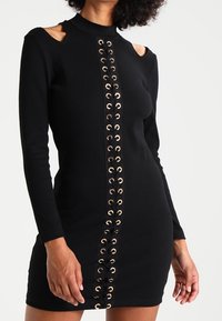 Svart långärmad klänning med utskurna axlar, med ett snörningsdetalj framtill med guldfärgad ögla. Åtsittande, tillverkad av stretchigt tyg.