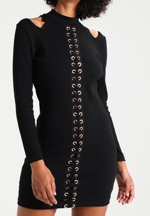 Schwarzes langärmliges Kleid mit ausgeschnittenen Schultern, das eine Schnürung mit goldfarbenen Ösen auf der Vorderseite zeigt. Figurbetont und aus dehnbarem Stoff.