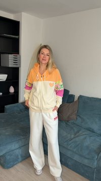 Kvinde med blondt hår står indendørs iført en creme- og orange hættetrøje med blomsterærmer og hvide bukser med vide ben ved siden af en blå sofa.