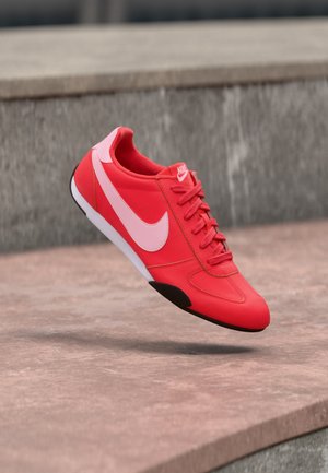 Κόκκινο αθλητικό παπούτσι Nike με ροζ λογότυπο και κορδόνια, αιωρούμενο πάνω από σκυρόδεμα κοντά σε πέτρινο τοίχο.