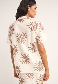 Camisa de manga corta en tela crema con patrones de sol marrón, que presenta un cuello clásico y un ajuste relajado. La vista trasera muestra la repetición del patrón.