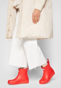 Bottes en caoutchouc rouges mi-mollets avec une finition lisse, dotées d'une languette à l'arrière et d'un logo blanc contrastant, associées à un manteau rembourré beige.