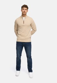 Beige zip-up tröja av stickad tyg, med hög krage och detaljer i brun dragkedja, kombinerad med mörkblå jeans och vita sneakers.