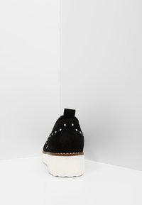 Zapatilla de ante negra con suela de plataforma blanca, que presenta tachuelas plateadas a lo largo de los lados y una lengüeta en la parte trasera.