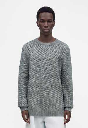 Jeune homme aux cheveux courts portant un pull en maille gris texturé et un pantalon clair sur un fond blanc uni.