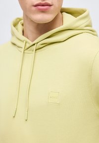 Κοντινή λήψη ενός ατόμου που φοράει φούτερ με κουκούλα ανοιχτού πράσινου χρώματος με κορδόνια και μικρό λογότυπο "BOSS" στο στήθος.