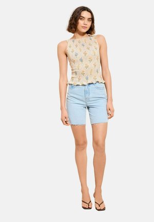 Kvinde iført en ærmeløs beige top med blomsterprint, lyseblå denimshorts og sorte klipklapper, der står mod en ensfarvet baggrund.