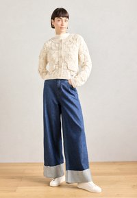 Häkelgestrickte cremefarbene Strickjacke mit Rundhalsausschnitt und Vorderknöpfen, kombiniert mit weiten blauen Jeanshosen mit kontrastierendem Saumdetail und weißen Sneakers.