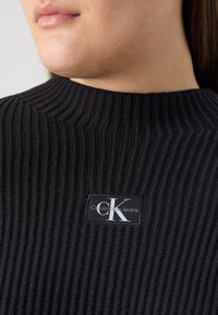 Pull en maille côtelée noir avec un col montant et une étiquette de marque arborant le logo "Calvin Klein Jeans". Détail de tissu texturé visible.