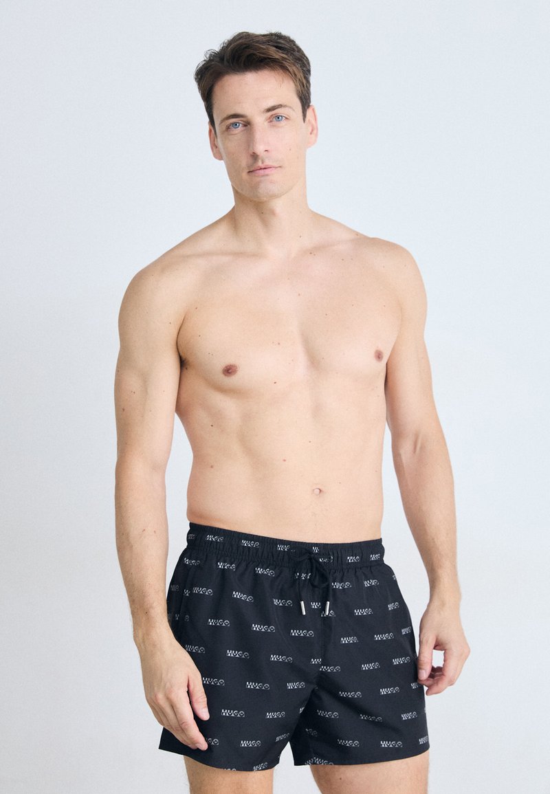 Svart svømmedraktshorts med et hvitt gjentatt logo-mønster, laget av lettvektsmateriale, med justerbar snøring i midjen.
