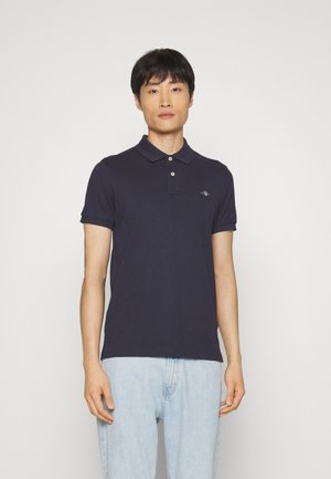 Marineblaues Poloshirt mit einem Kragen mit zwei Knöpfen, kurzen Ärmeln und einem kleinen gestickten Logo auf der linken Brust, kombiniert mit hellblauen Denimjeans.