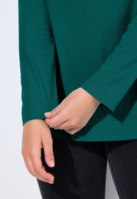 Tyrkysový top s dlouhými rukávy, bavlněný materiál, kulatý výstřih, přiléhavé rukávy, jemné detaily v prošívání, nošený s tmavými džíny.