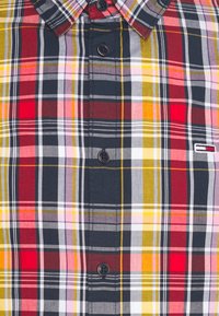 Primo piano di una camicia a quadri multicolore con quadrati rossi, gialli, navy, rosa e bianchi, bottoni neri e un piccolo logo rettangolare.