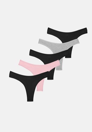 Jeune femme portant un débardeur noir court et un sous-vêtement assorti à taille haute, debout devant un fond clair uni, regardant vers l'avant.