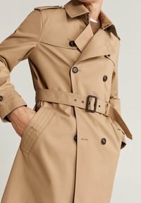 Beżowy trencz z dwurzędowym zapięciem, paskiem w talii, dużymi czarnymi guzikami i naramiennikami. Materiał o fakturze, klasyczny styl.