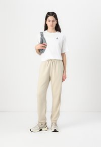 Hvit t-skjorte med kortermet stilling og svart logo, beige joggebukser og lys grå sko. Holder en grå skulderveske. Enkelt, uformelt stil.