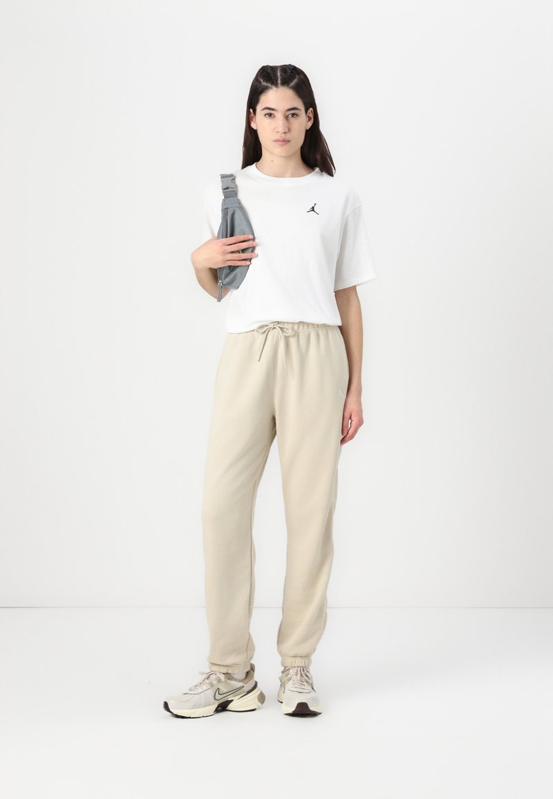 Weißes T-Shirt mit kurzen Ärmeln und schwarzem Logo, beiger Sweatpants und hellgrauen Sneakers. Hält eine graue Umhängetasche. Schlichter, lässiger Stil.