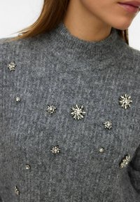 Grijze gebreide trui met een gestructureerd patroon en een hoge hals, voorzien van meerdere decoratieve bloemrijke versieringen in zilverkleurige tinten.