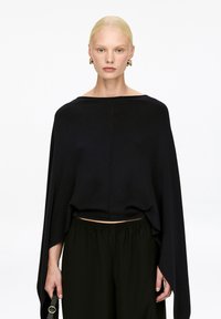 Schwarzes, kurzes Oberteil mit einem lockeren, drapierten Design und weiten Ärmeln, kombiniert mit schwarzen Hose mit elastischem Bund. Glattes Material mit minimaler Naht.