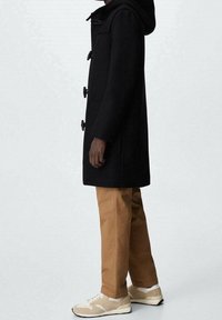 Manteau duffle en laine noire avec fermetures à boutons à bascule et capuche, associé à un pantalon beige et des baskets crème au design texturé.