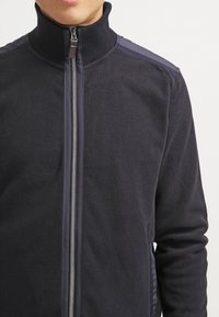 Giacca in pile blu navale con colletto alto, chiusura con zip e dettagli in tessuto testurizzato sulle spalle. Presenta una zip liscia.