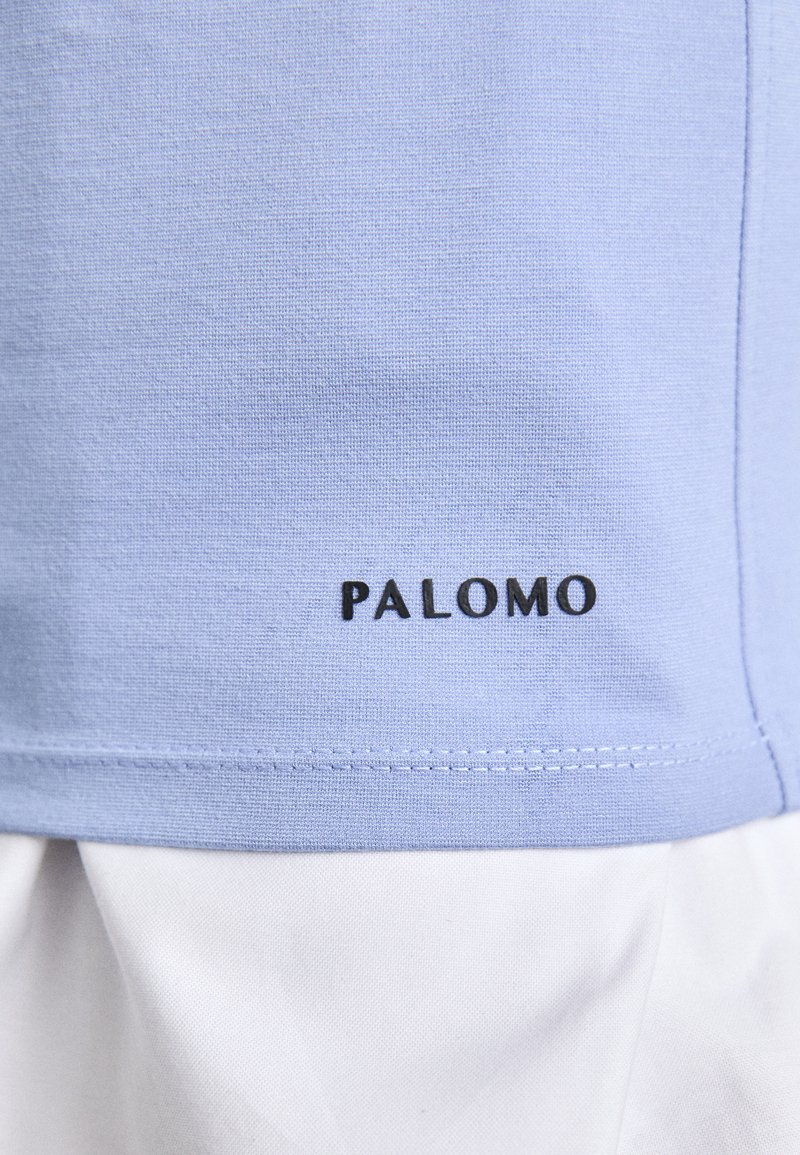 Lichtblauwe katoenen shirt met witte stof eronder, voorzien van een gestikte zoom en het woord "PALOMO" in zwart aan de onderkant.