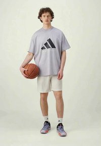 Joven de pie con un balón de baloncesto, vistiendo una camiseta de baloncesto gris de Adidas, pantalones cortos claros, calcetines blancos de Adidas y zapatillas grises.