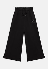 Breda svarta sweatpants gjorda av mjukt material, med elastisk midja, sidofickor och en liten vit logotyp på vänster sida.