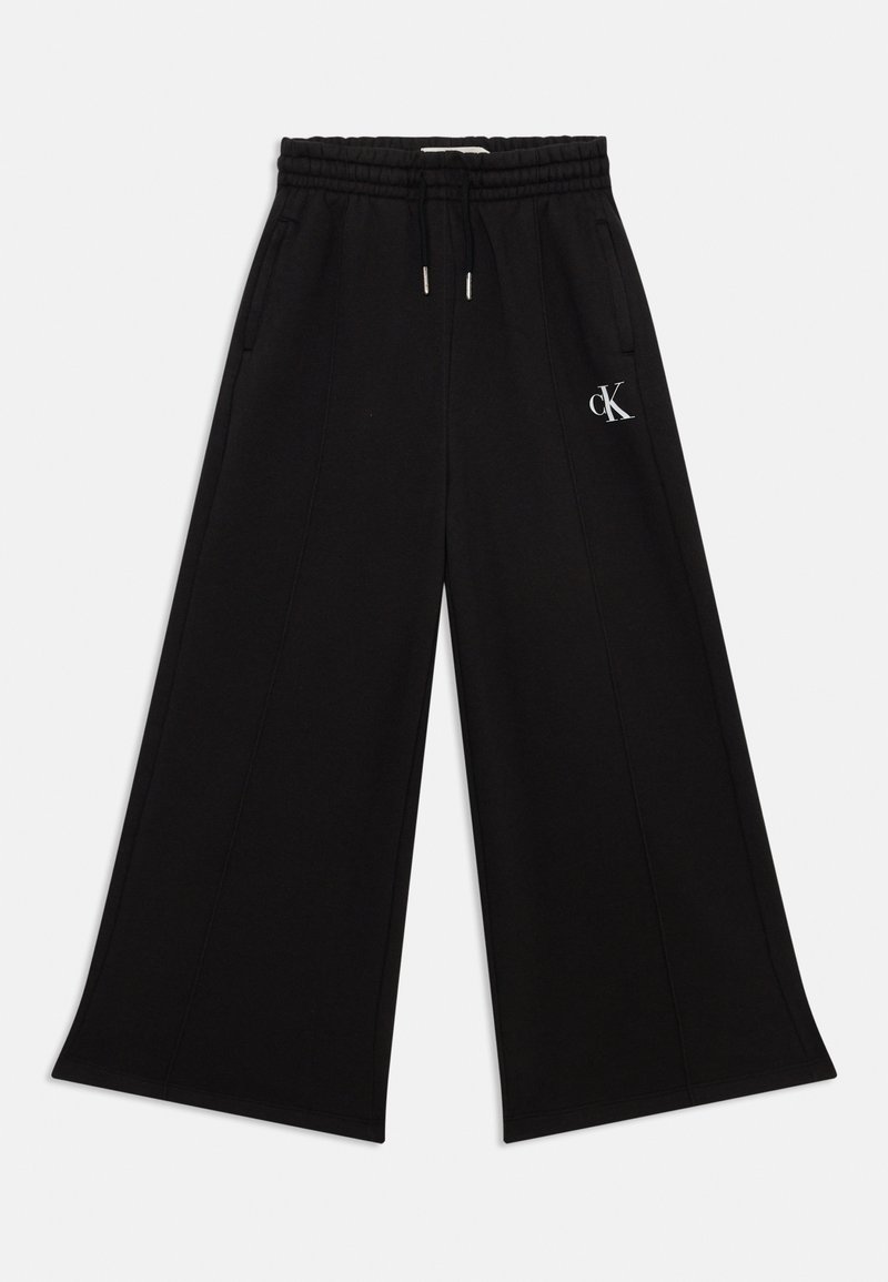 Breda svarta sweatpants gjorda av mjukt material, med elastisk midja, sidofickor och en liten vit logotyp på vänster sida.