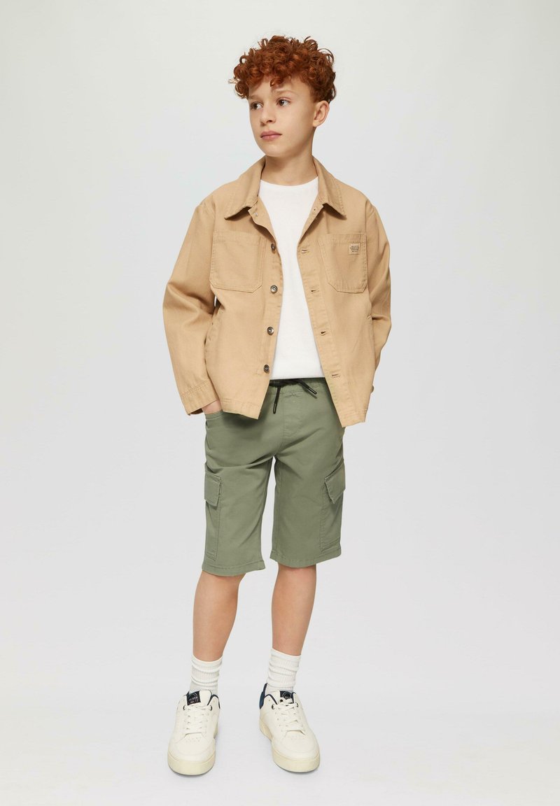 Hellbraunes Hemd mit Knöpfen, weißes T-Shirt, grüne Cargo-Shorts, weiße Socken und Turnschuhe. Lockige Haare, steht mit den Händen in den Taschen.