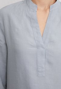 Ljusblå linblus med mandarin-krage, med halvknappar och subtil textur. Tyget verkar mjukt och andningsbart.