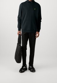 Sudadera negra con bolsillo tipo canguro y un pequeño logo amarillo, combinada con pantalones negros ajustados y un bolso de mano acolchado negro. Zapatillas negras con toques blancos.