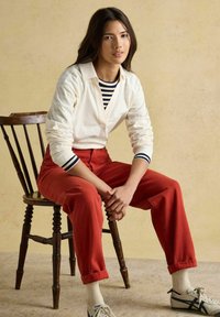 Noor naine istub puidust toolil, kandes valget pikka varrukat särki triibulise pluusi üle, punaseid pükse, valgeid sokke ja valgeid spordijalanõusid, millel on tumesinised triibud.